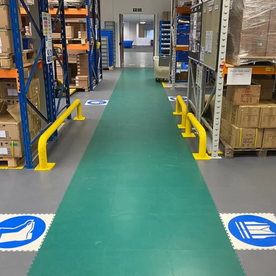 Image of a green walkway in a warehouse, with floor signage on either direction symbolsing PPE requirements
