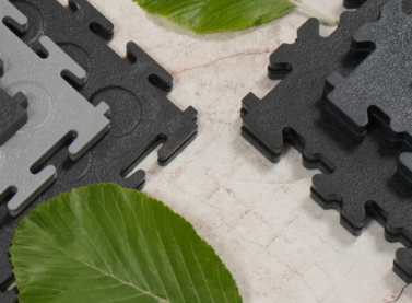Une gamme de dalles de sol emboîtables recyclées Ecotile posées sur un sol en béton brut, accompagnées de feuilles vertes symbolisant la durabilité et le caractère écologique du produit