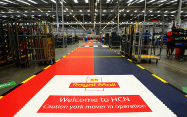 Revêtement de sol d'un entrepôt de Royal Mail avec pose d'Ecotile sur les allées, associant des dalles colorées au logo de la marque et à des messages d'avertissement destinés aux chariots élévateurs en service.